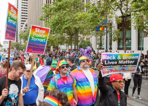 Image from a Pride protest, containing a crowd of people dressed in rainbow colours carrying rainbow placards